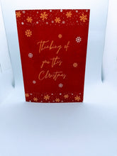 Load image into Gallery viewer, The Thinking of You Christmas Card has an festive crimson background with a gold snowflake and white snow motif. The message in gold reads &quot;Thinking of you this Christmas&quot;. 
